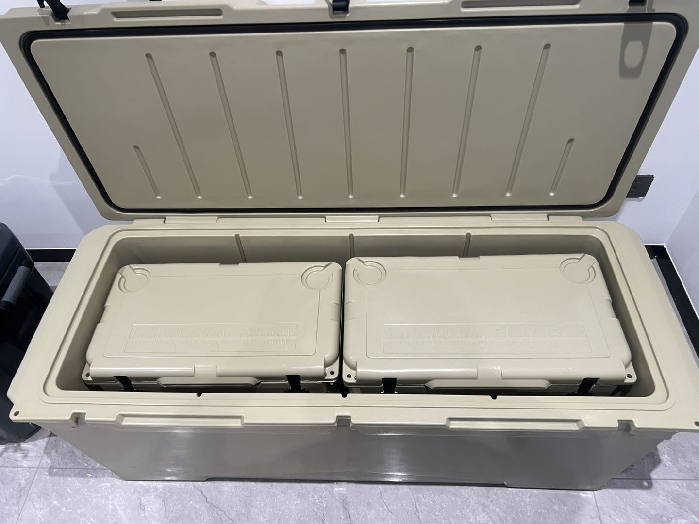 A large beige insulated cooler box with its lid open, showing two smaller matching cooler boxes nested inside on a gray tiled floor.